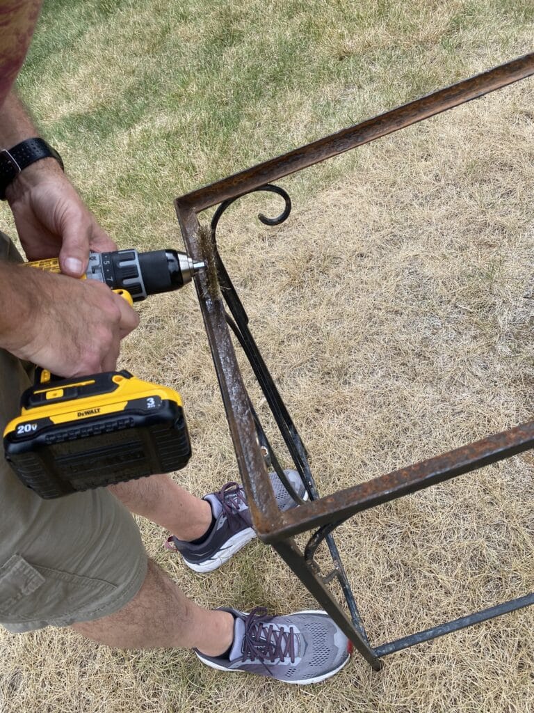 Man sanding rusted wrought iron table with wire brush attached to power drill