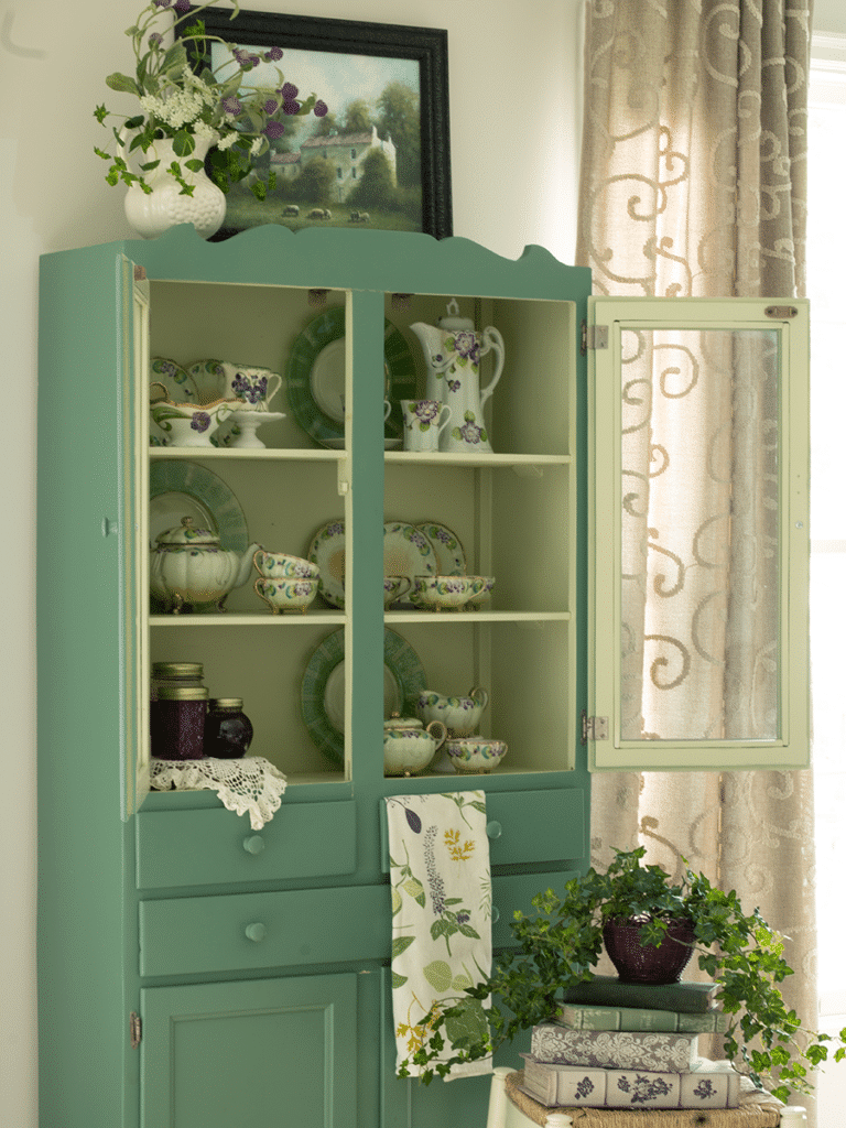 A vintage kitchen cabinet, painted green housing a purple and green teaset