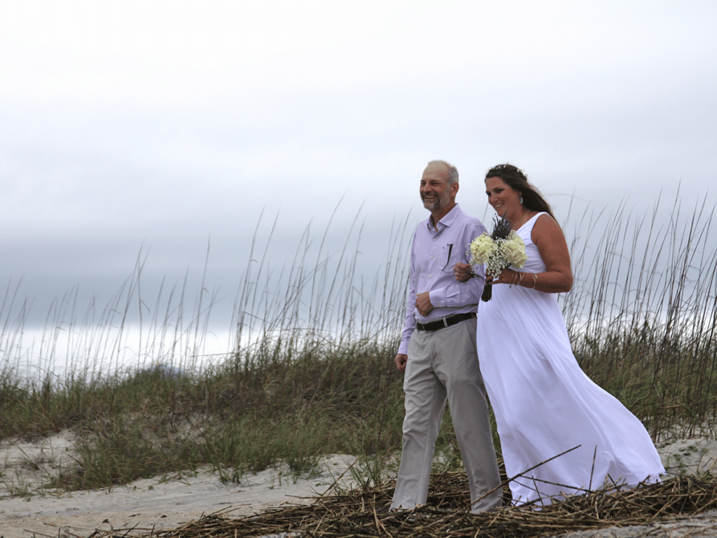 A father of the bride walks his daughter across beach dunes