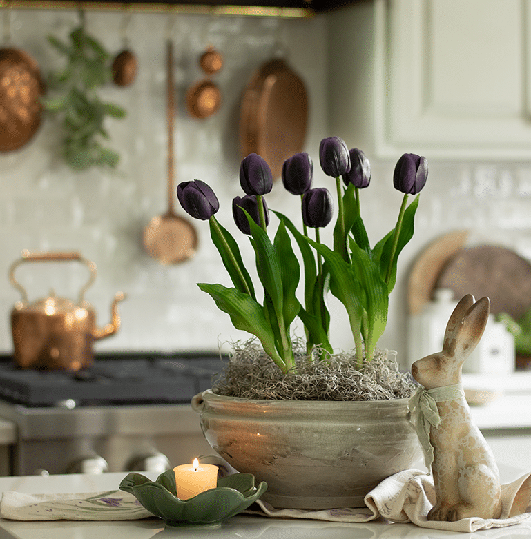 A cache pot filled with faux tulips on a kitchen island with a copper pot rail in the background.